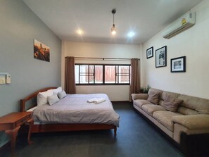 Un dormitorio con cama, mesita de noche de madera, sofá beige, ventana con cortinas y aire acondicionado instalado en la pared.