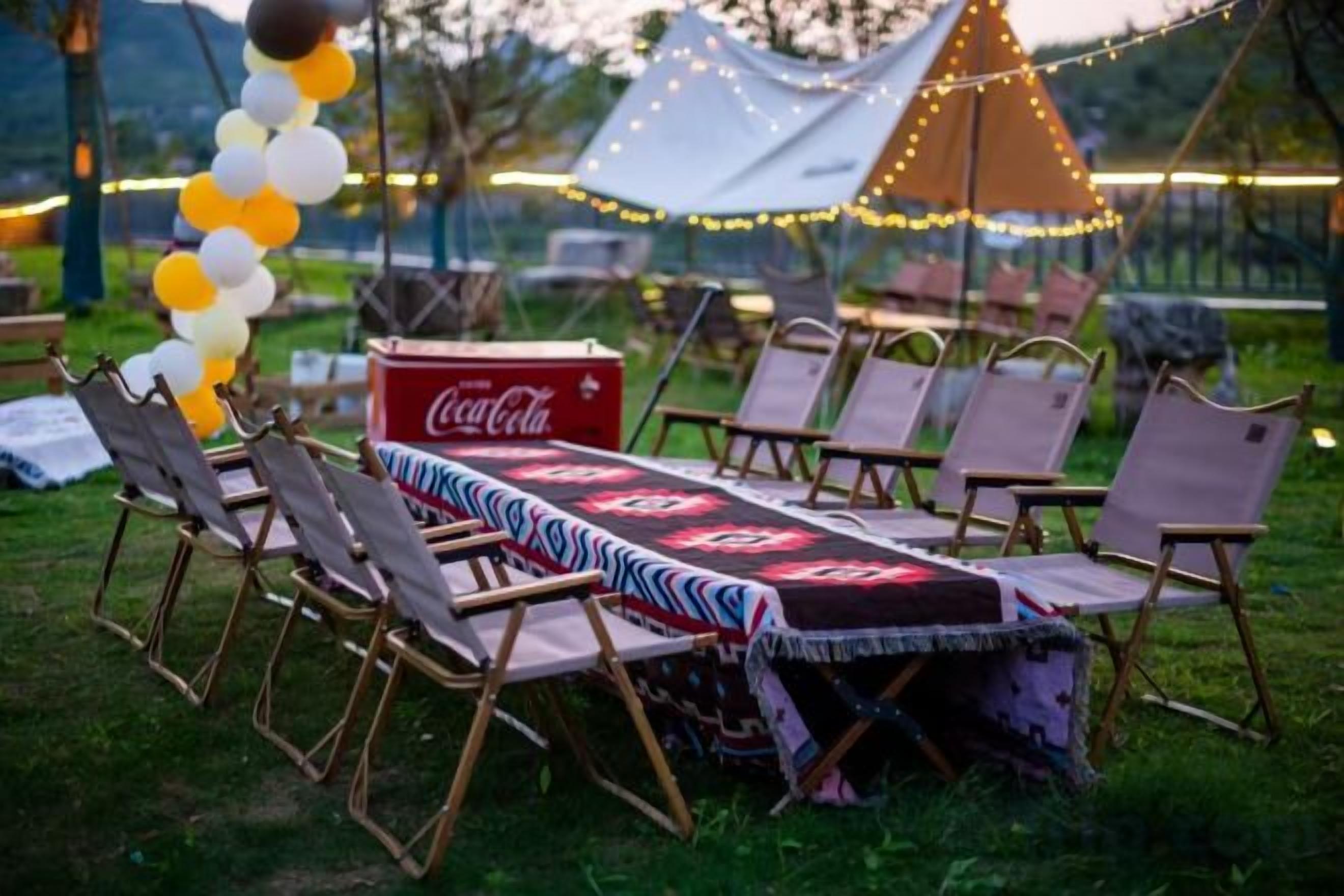 Ulkoilma-alue, jossa on pöytä kuudelle henkilölle, pöydän ympärillä tuoleja ja pöydällä Coca-Cola-juomajääkaappi.
