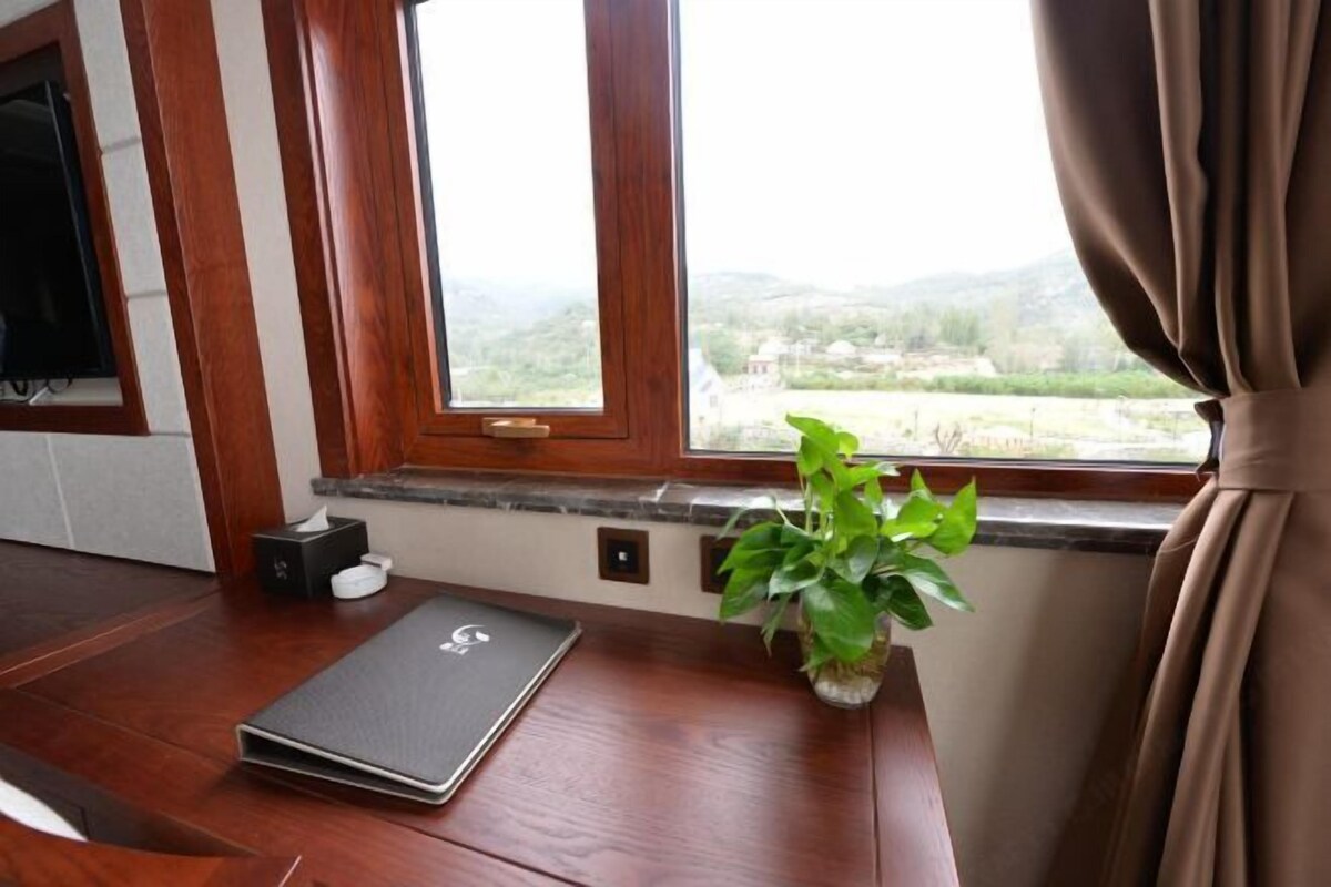 A room with a wooden desk, a laptop, a plant, and a window with a view of the countryside.