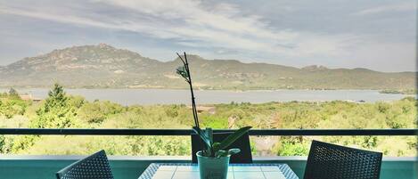 A balcony with a table and chairs, overlooking a mountainous landscape and a body of water.