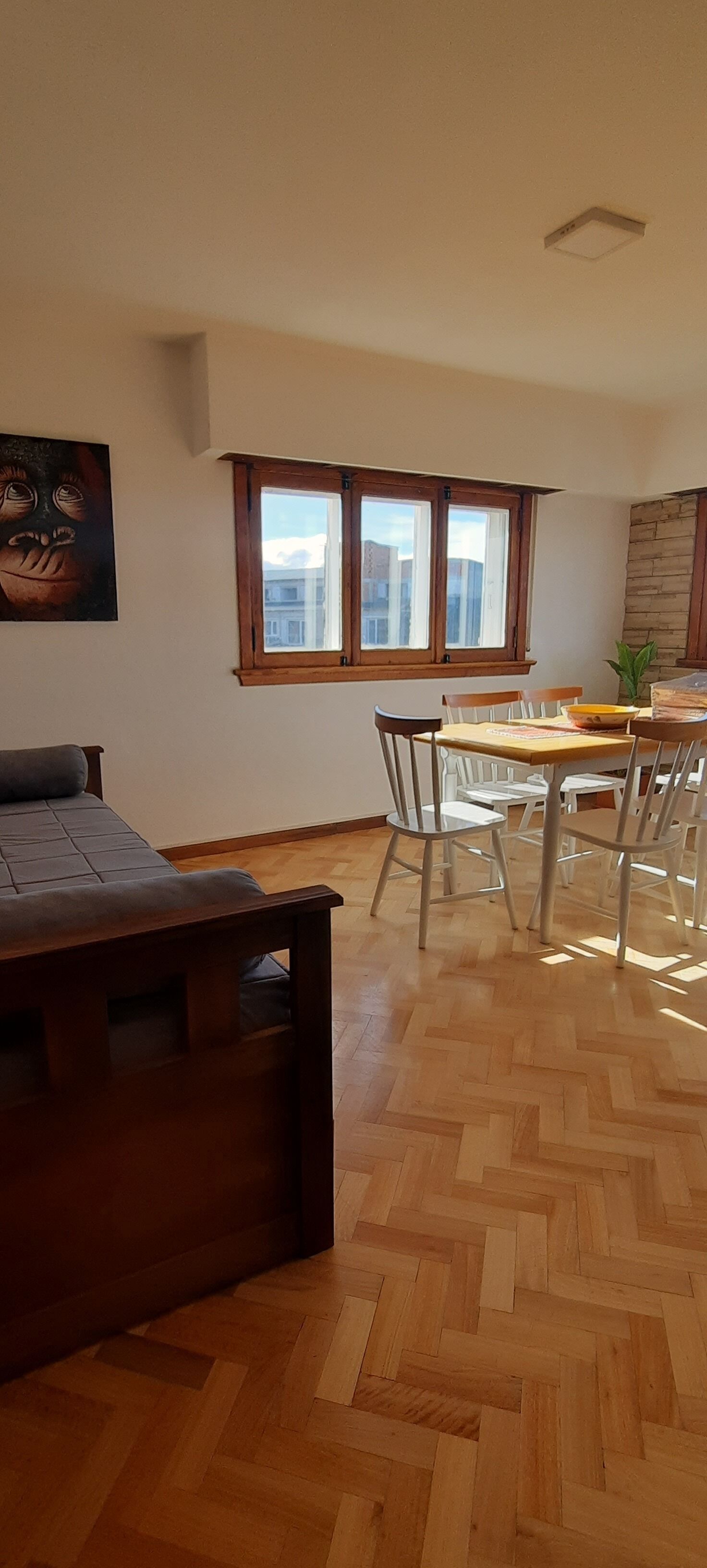 Una habitación con piso de madera en parquete, una mesa de comedor con sillas, un ventanal con vista a edificios y un cuadro en la pared.