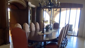 Ein Esszimmer mit einem großen runden Tisch, mehreren gepolsterten Stühlen, einer Chandelierleuchtre und Blick durch große Fenster.