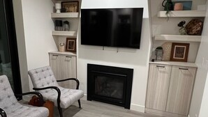 A modern living room with a flat-screen TV mounted above a fireplace, grey upholstered chairs, and built-in shelving with decorative items. - Steps away from the slopes!  Recently built Scandinavian Cabin with Jacuzzi! (Beaver)