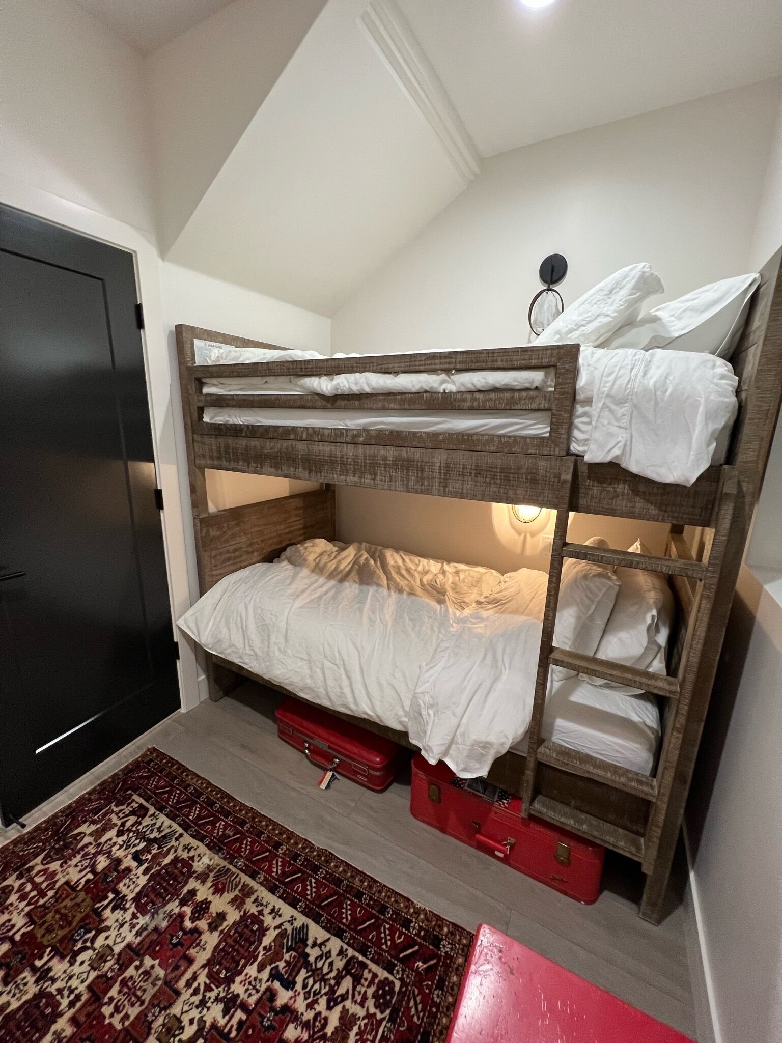 Un letto a castello con struttura in legno, biancheria bianca, una valigia rossa sotto il letto inferiore e un tappeto decorato sul pavimento.
