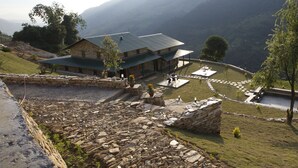 Un edificio de piedra con techo verde, rodeado por un muro de piedra y un área con césped y algunos árboles.