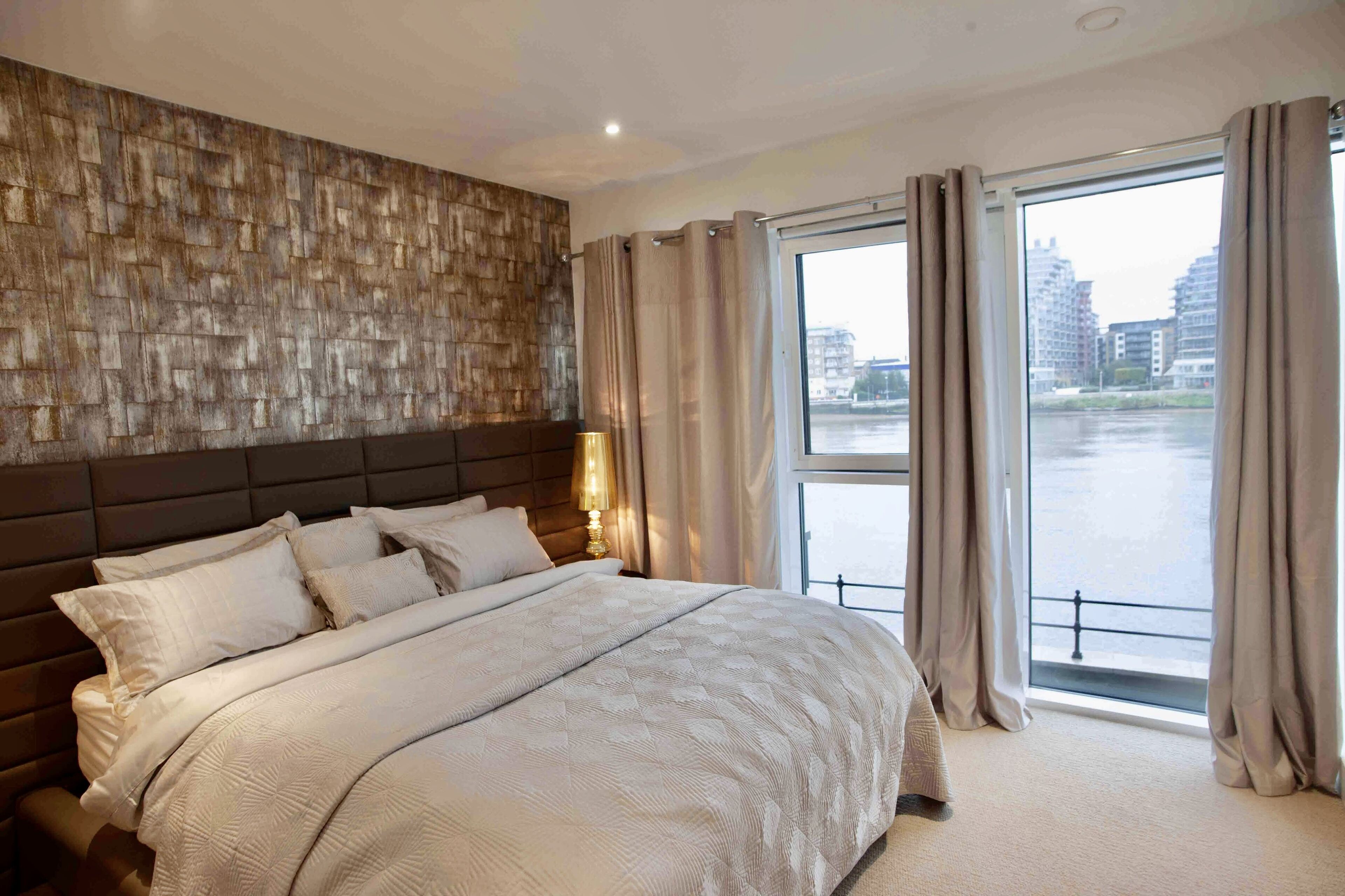 Ein Hotelzimmer mit einem großen Bett, einem Kopfteil mit einer strukturierten Wand und Blick auf ein Gewässer durch große Fenster.
