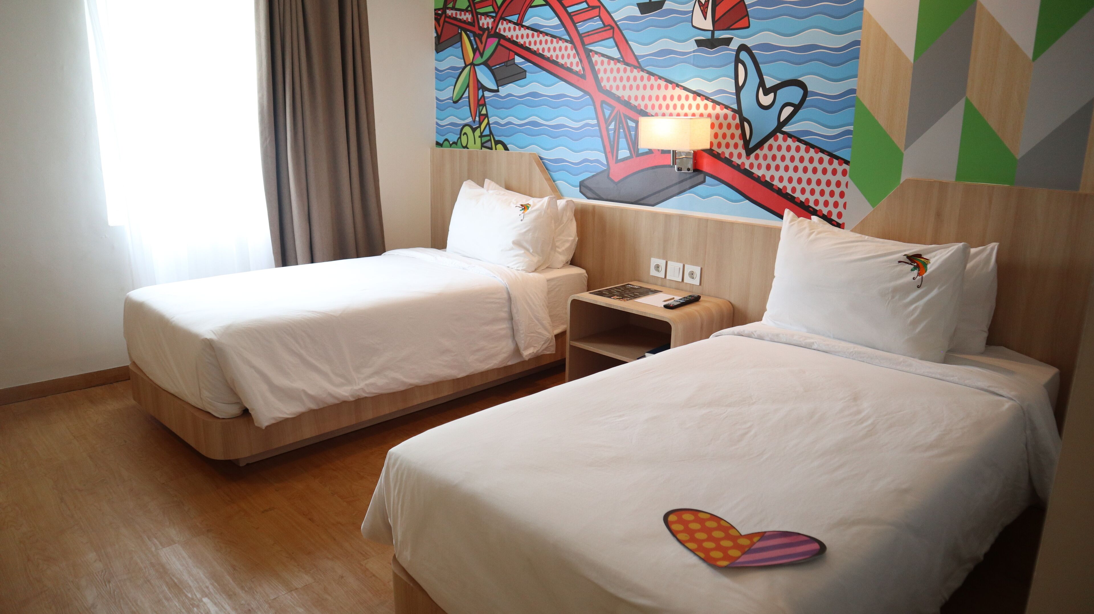 A hotel room with two single beds, a nightstand, a lamp, and a colorful mural on the wall.