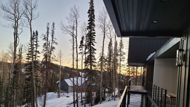 Une terrasse en bois recouverte de neige, donnant sur une zone boisée avec une maison en arrière-plan.