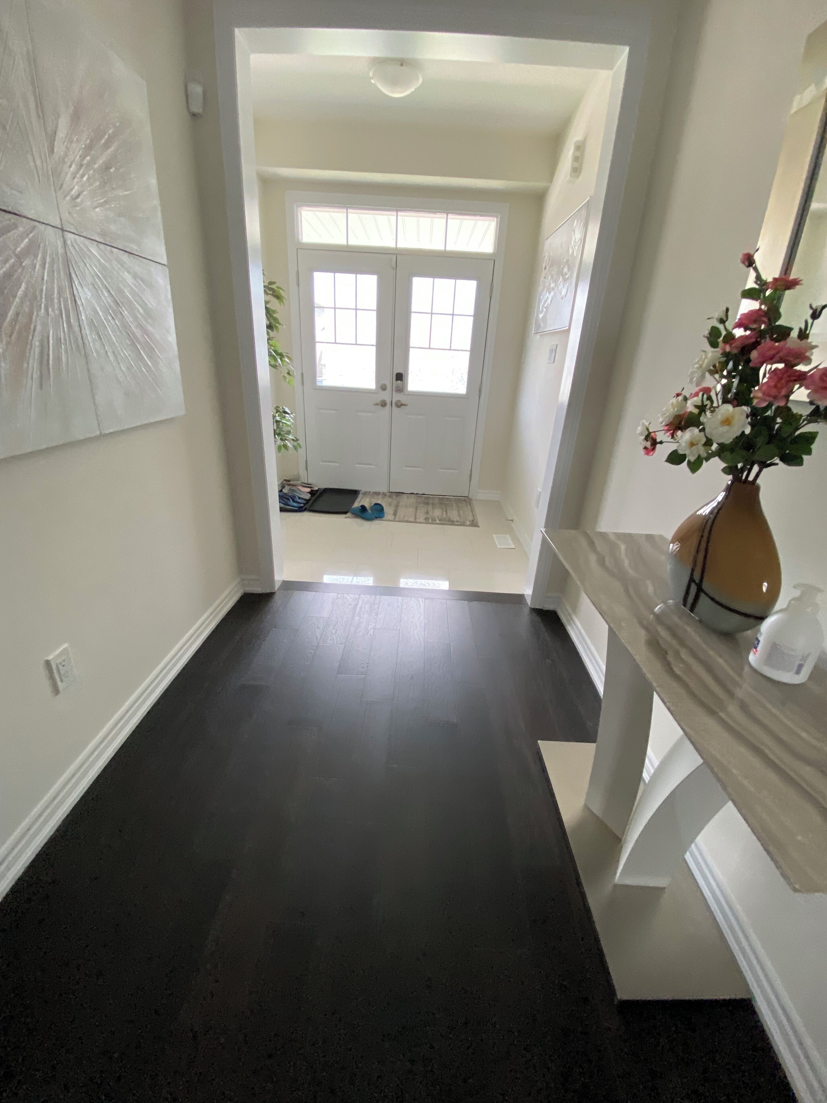 A hallway with a dark floor, a white door with windows, and a vase with flowers on a white console table.