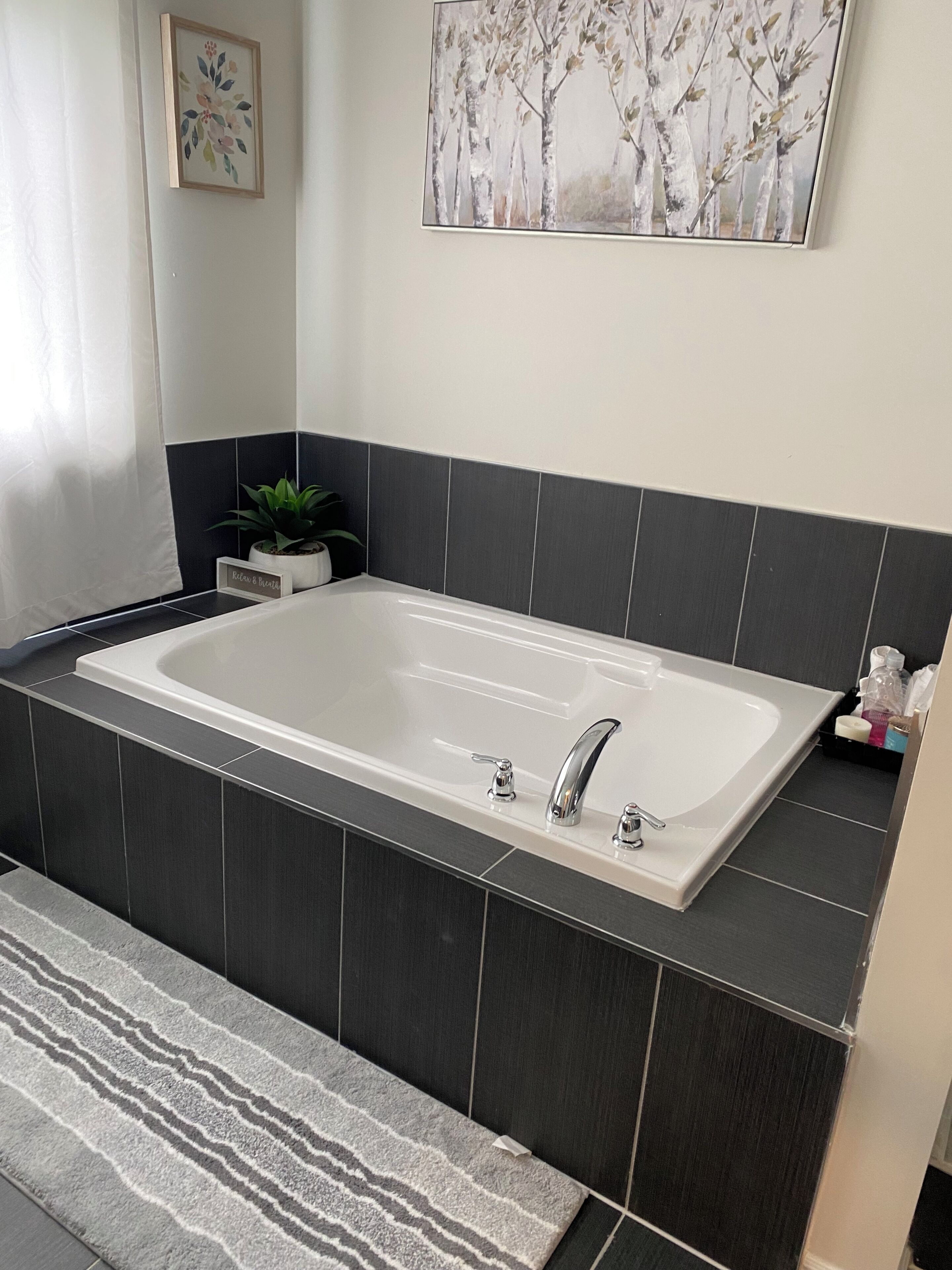 A modern bathroom with a large bathtub, dark tiled walls, a small potted plant, and a framed artwork above the tub.