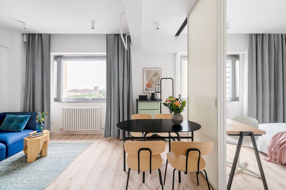 Een moderne woonkamer met een bank, een eettafel met stoelen, een klein bijzettafeltje, een radiator en een raam met gordijnen.