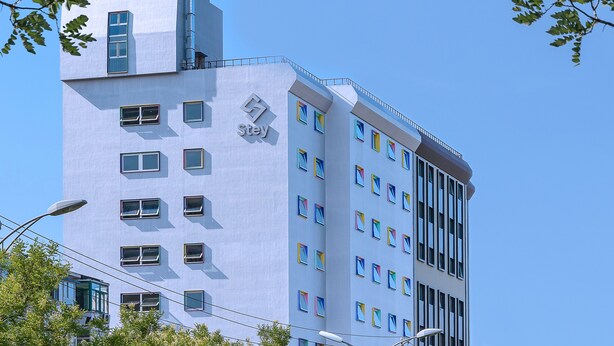 Un bâtiment moderne arborant un logo « stey » bien visible, doté de nombreuses fenêtres et se dressant sur fond d’un ciel bleu clair.