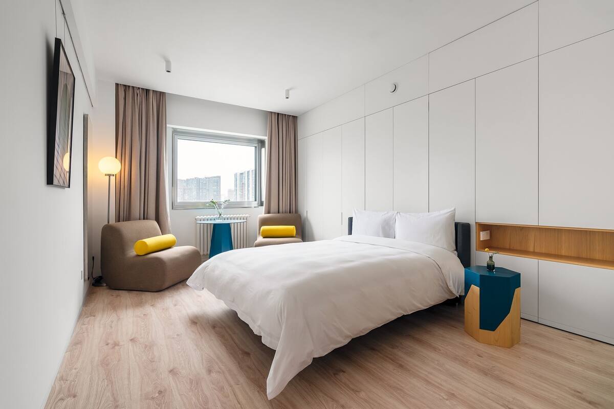 Een moderne hotelkamer met een groot bed, twee fauteuils, een bijzettafeltje en een raam met uitzicht op de stad.