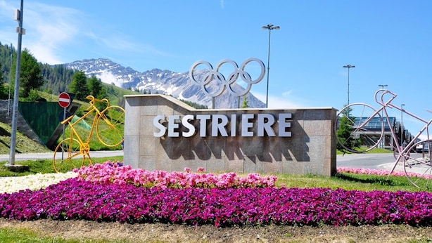 En skylt med texten ”SESTRIERE” och de olympiska ringarna, omgiven av blommor och med ett berg i bakgrunden.
