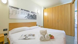 A hotel room with a large bed, a wooden wardrobe, a painting on the wall, and a bedside table with a lamp. - Teleo 115 Ski In - Ski Out 50m, Sestriere, Italy (Sestriere)