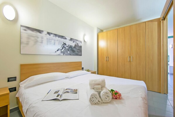 A hotel room with a large bed, a wooden wardrobe, a painting on the wall, and a bedside table with a lamp.
