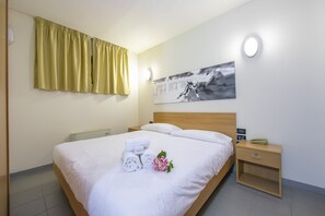 A neatly made bed with white linens, a bedside table with a lamp, a wall-mounted artwork, and a window with yellow curtains.