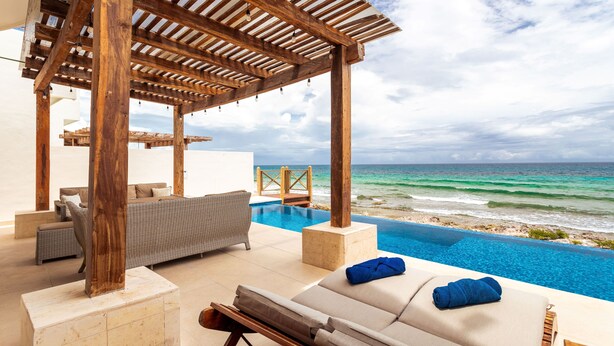 Een overloopzwembad met uitzicht op de oceaan, een houten pergola en een loungeruimte met kussens.