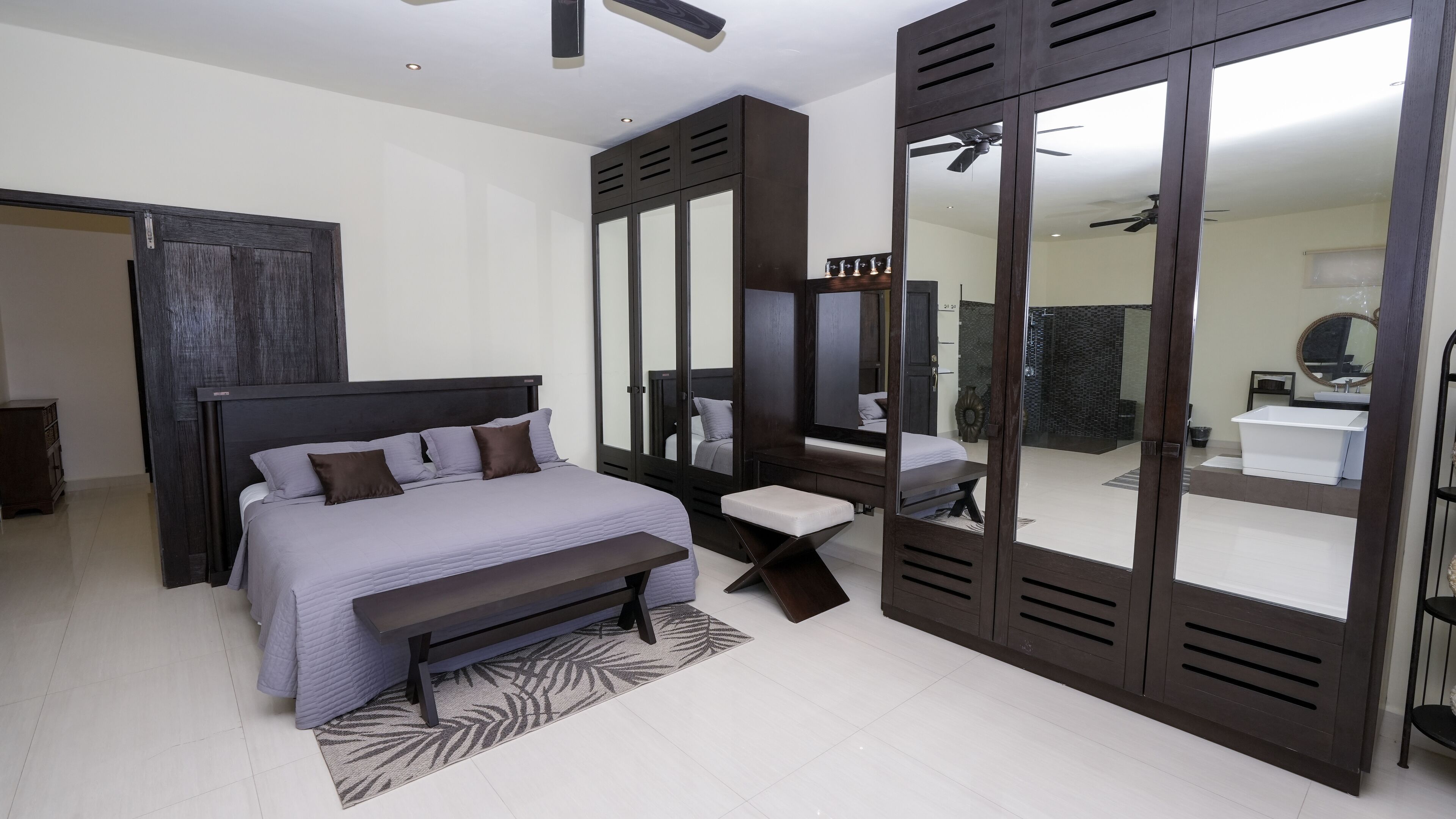 A modern bedroom with a large bed, a bedside table, a bench, a wardrobe with a mirror, and a view into a bathroom with a bathtub.