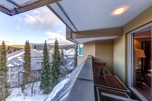 Un balcon donnant sur des maisons et des arbres couverts de neige, une table avec des chaises et une fenêtre avec une lampe.