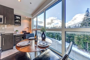 A modern kitchen with dark wood cabinets, a dining table set for four, and a large window offering a view of a snowy landscape.