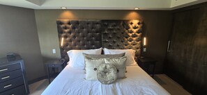 Une chambre à coucher avec une tête de lit capitonnée, un lit recouvert de draps blancs, plusieurs oreillers, des tables de chevet avec des lampes et une commode.