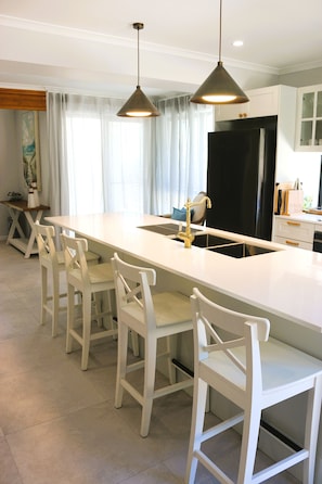 Una cocina moderna con una isla central, taburetes blancos de bar, iluminación colgante y un refrigerador grande.