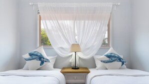 Un dormitorio con dos camas, una mesita de noche con lámpara y una ventana con cortinas traslúcidas.