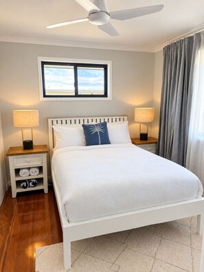 A bedroom with a bed, two bedside tables, a ceiling fan, a window with curtains, and a view of the sky. - Lovely Waterfront Beach House, Pet-Friendly (Buddina)