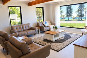 A modern living room with leather sofas, a wooden coffee table, and a view of a tropical landscape through large windows. - Lovely Waterfront Beach House, Pet-Friendly (Buddina)