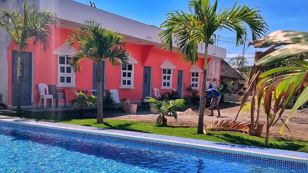 Une piscine à l’eau d’un bleu cristallin, entourée d’un bâtiment rouge aux fenêtres et aux portes à cadre blanc, et de palmiers.