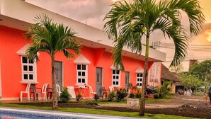 Une piscine à l’eau d’un bleu cristallin, entourée d’un bâtiment rouge aux fenêtres et aux portes blanches, et de palmiers.