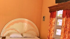 Une chambre à coucher comprenant un lit, une table de chevet, un téléviseur fixé au mur et une fenêtre agrémentée de rideaux à motifs.