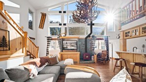 A cozy living room with a fireplace, wooden furniture, and a large window offering a view of the outdoors. - Spacious, Luxury, Picturesque "Mountain High Manor"- Ski Resort Town! (Wrightwood)