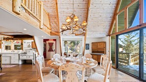 A spacious dining area with a high ceiling and a chandelier, wooden chairs, and a large window offering a view of the outdoors. - Spacious, Luxury, Picturesque "Mountain High Manor"- Ski Resort Town! (Wrightwood)