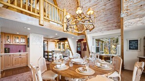 A dining area with a wooden table set for six, surrounded by white wicker chairs. A chandelier with antlers hangs above the table. - Spacious, Luxury, Picturesque "Mountain High Manor"- Ski Resort Town! (Wrightwood)