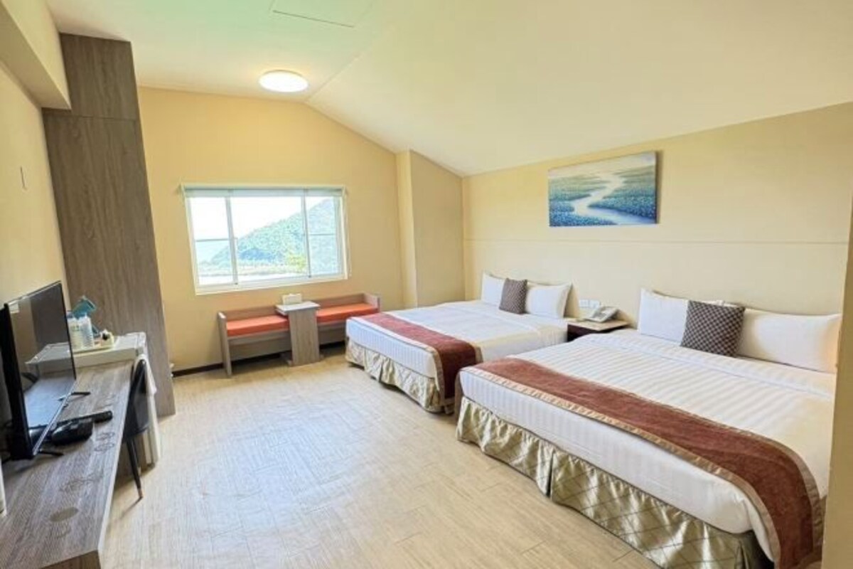 Quarto de hotel com duas camas, uma escrivaninha com computador, janela com vista para colinas e um quadro na parede.