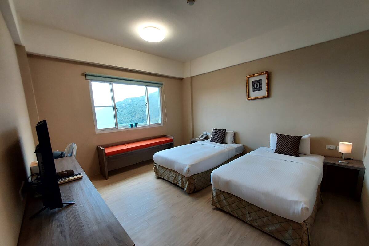 Ein Hotelzimmer mit zwei Einzelbetten, einem Schreibtisch mit Computer, einem Fenster mit Bergblick und einem Nachttisch mit Lampe.