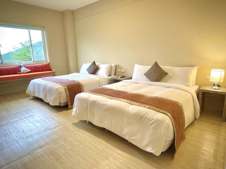 A hotel room with two beds, a window seat with a red cushion, and a lamp on the nightstand.