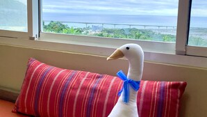 Ein weiches Entenspielzeug mit blauem Band auf einem Fensterbrett, ein gestreiftes Kissen und Blick auf das Meer und eine Brücke.