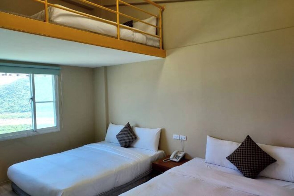 Quarto de hotel com duas camas de solteiro, uma cama beliche em cima, telefone ao lado da cama e janela com vista para o verde.