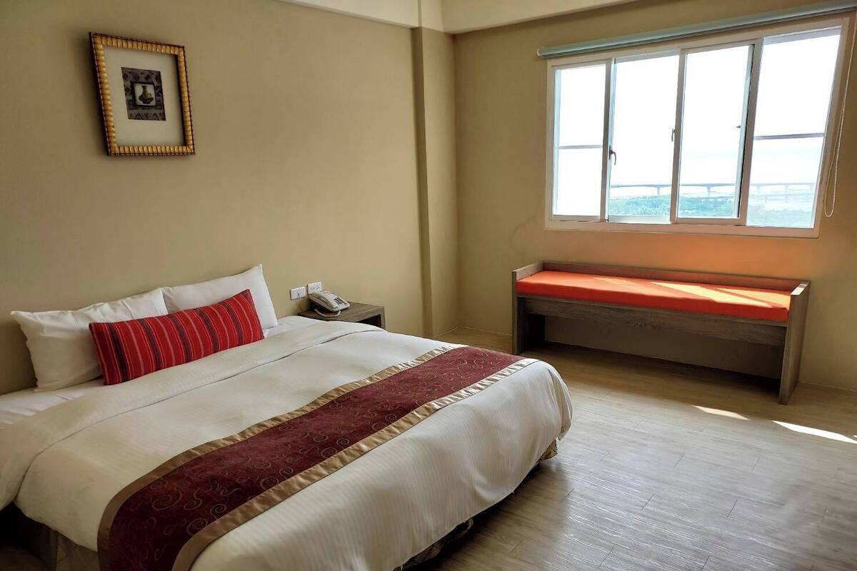 Ein Hotelzimmer mit einem großen Bett, einem Nachttisch mit Telefon, einem Fenster mit Sitzbank und einem gerahmten Bild an der Wand.