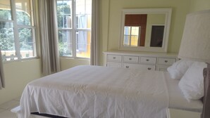 Ein ordentlich bezogenes Bett in einem Zimmer mit einem großen Fenster, einer Kommode mit mehreren Schubladen und einem Spiegel darüber.
