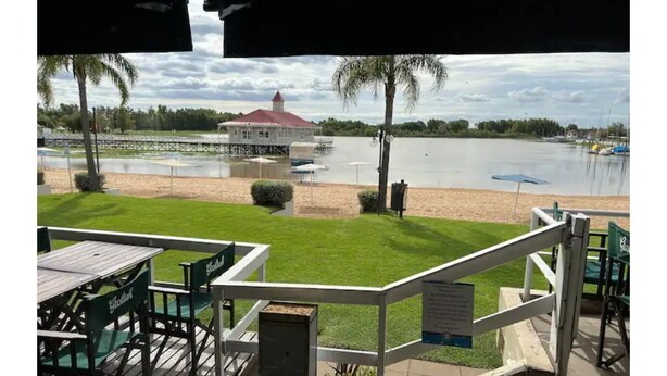 Une terrasse avec des chaises vertes et une table, donnant sur un lac, avec un bâtiment et des palmiers en arrière-plan.