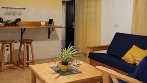 Un espace de vie compact comprenant une table à manger en bois, deux tabourets en bois, un canapé bleu avec un coussin jaune, une plante en pot et un luminaire suspendu.