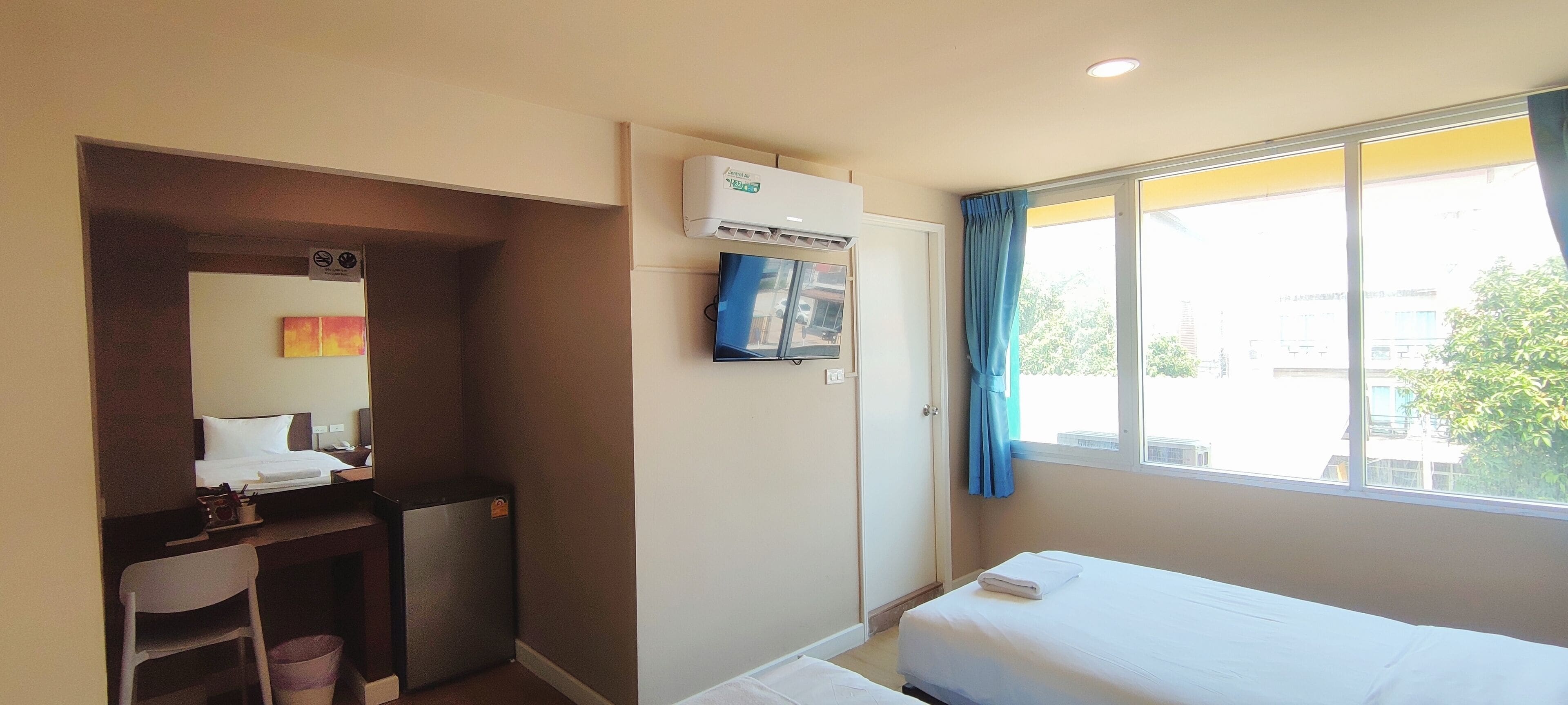 Une chambre d’hôtel équipée d’un lit, d’un bureau, d’une chaise, d’un petit réfrigérateur, d’un climatiseur, d’une télévision et d’une fenêtre avec des rideaux.
