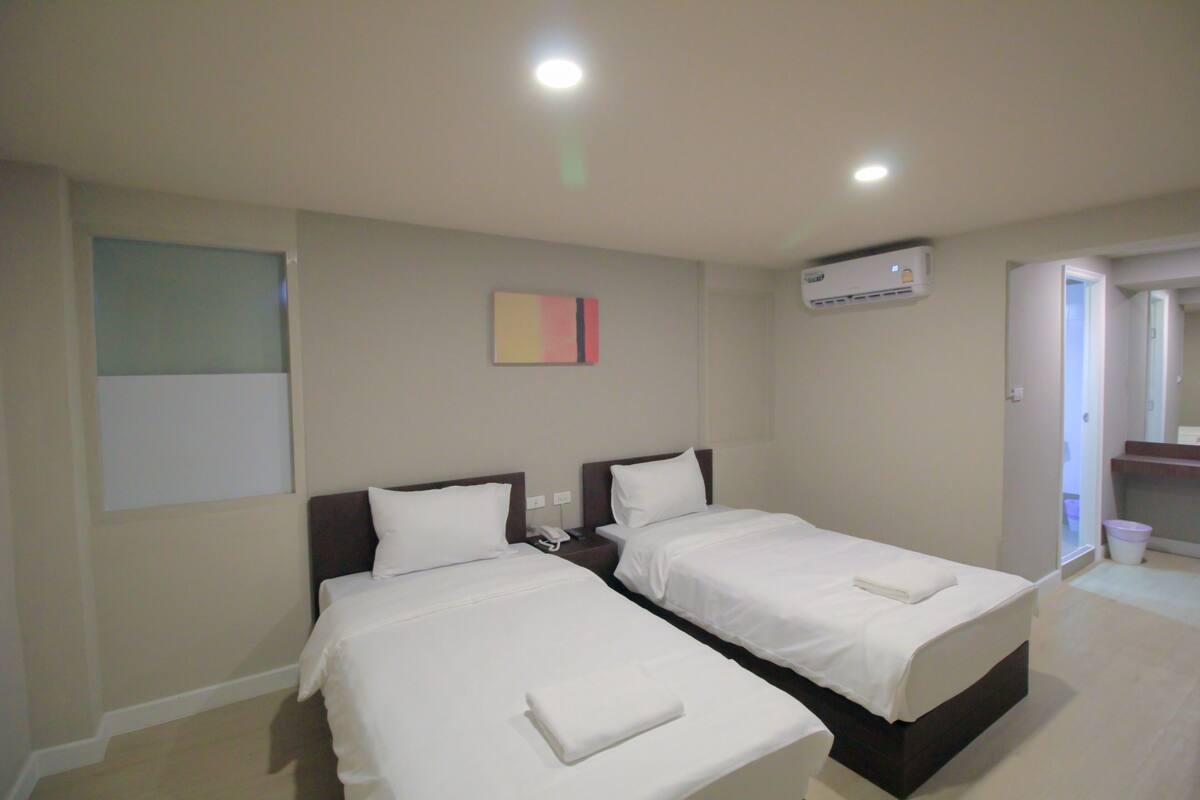 A hotel room with two beds, a nightstand, an air conditioning unit, and a painting on the wall.