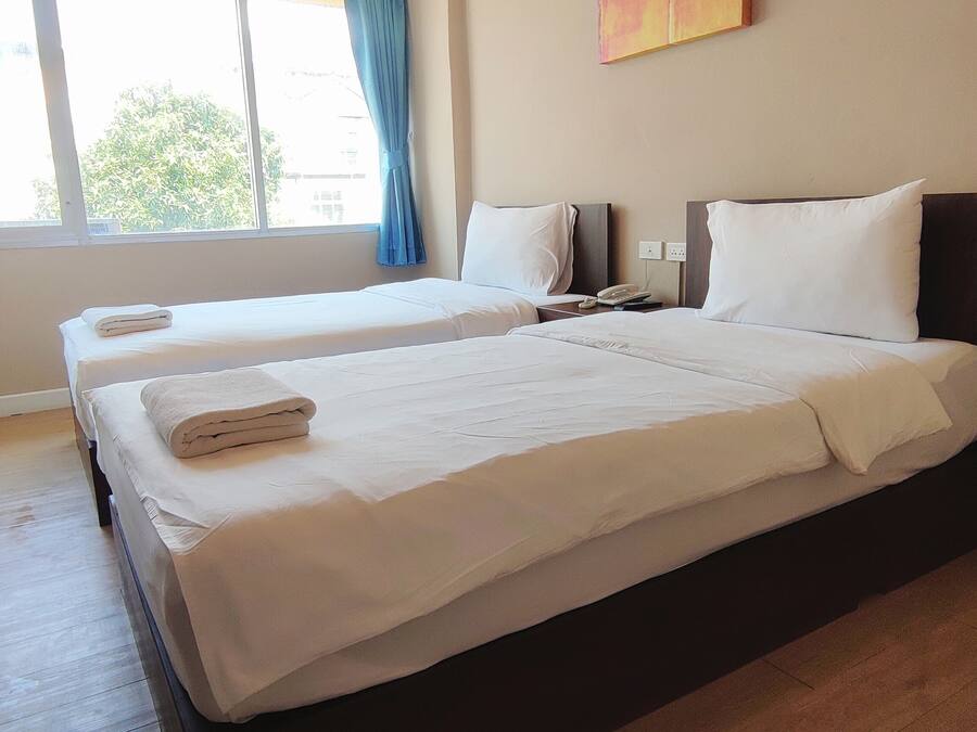 Ein Hotelzimmer mit zwei Betten, einem fensterbelaubten Fenster, einem Nachttisch mit Telefon und einem an der Wand befestigten Lichtschalter.