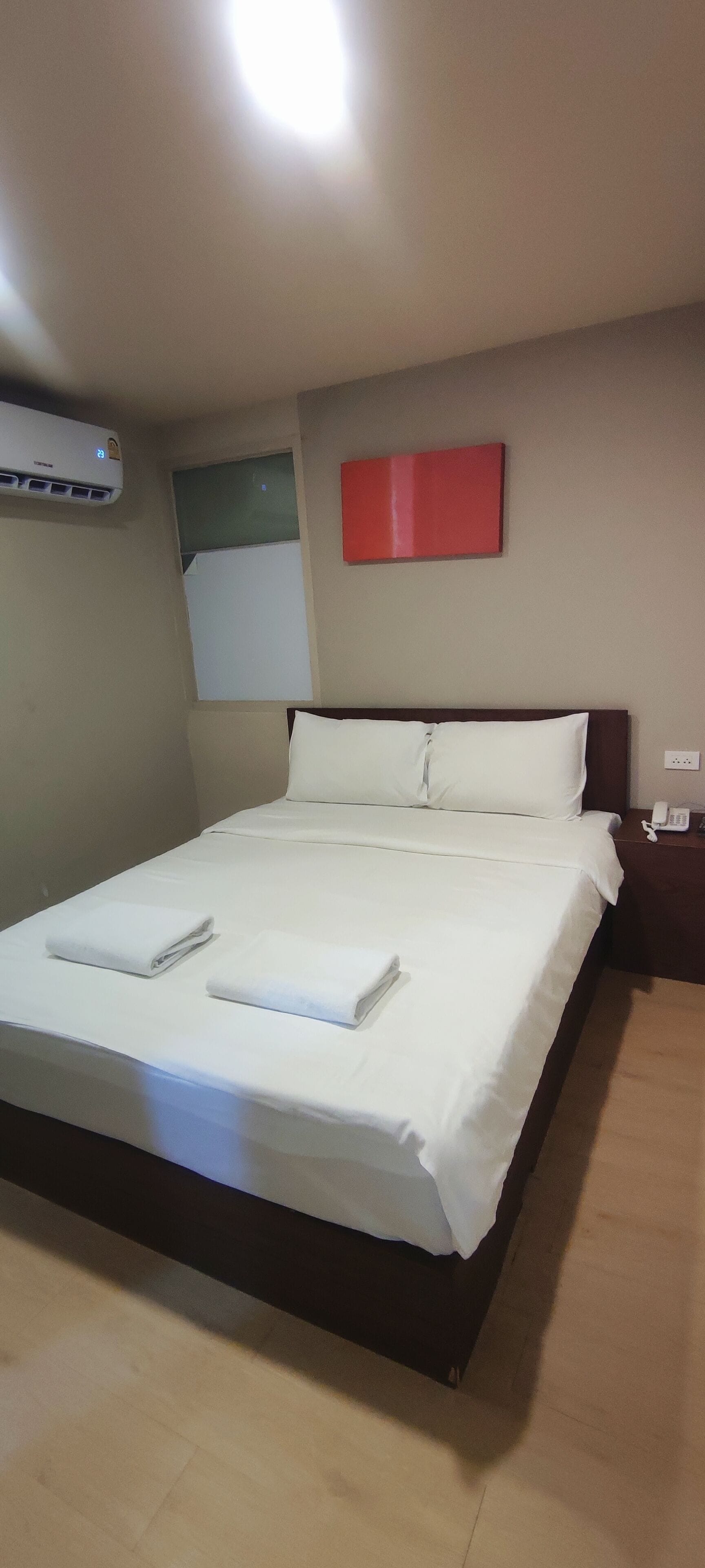 Una cama bien hecha con sábanas blancas y dos toallas dobladas, un adorno rojo en la pared superior y una unidad de aire acondicionado montada en la pared a la izquierda.