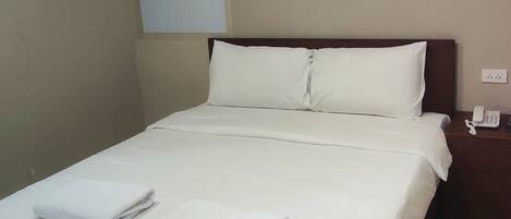 Uma cama bem arrumada com lençóis brancos e duas toalhas dobradas, uma decoração vermelha na parede acima dela, e um ar condicionado fixado na parede à esquerda.